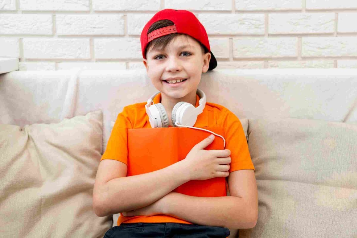 Smiling boy in an orange shirt and red cap holding a bright orange book with headphones around his neck, representing confidence and readiness to pass the 11 Plus English exam.